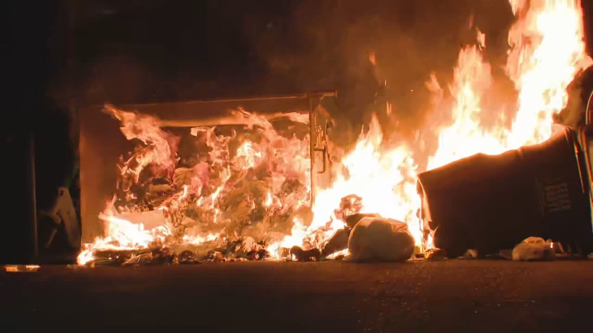 One of the dumpsters that was flipped has now been set on fire. @komonewsThe group is now blocking Stevens Way. Members of the group are intermittently blocking press cameras with umbrellas. No indications the people occupying the building have left.