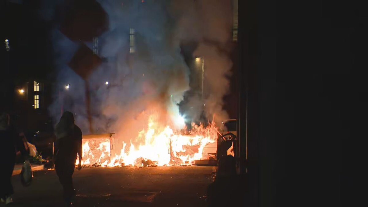 One of the dumpsters that was flipped has now been set on fire. @komonewsThe group is now blocking Stevens Way. Members of the group are intermittently blocking press cameras with umbrellas. No indications the people occupying the building have left. 