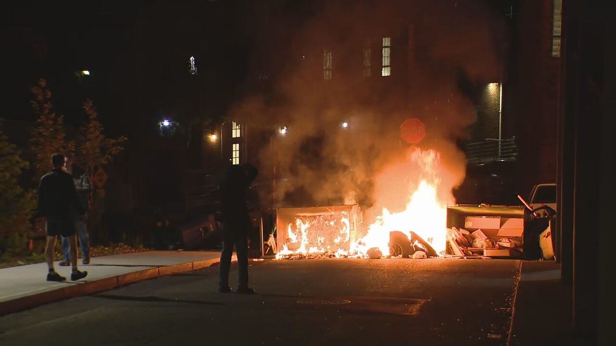 One of the dumpsters that was flipped has now been set on fire. @komonewsThe group is now blocking Stevens Way. Members of the group are intermittently blocking press cameras with umbrellas. No indications the people occupying the building have left. 
